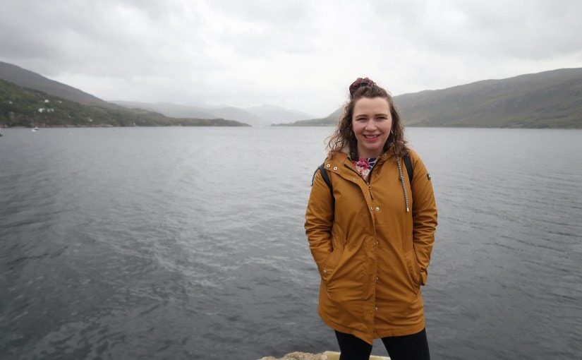 Picture shows Abi standing infront of a large lake. There are low mountains in the background and the sky is overcast and gloomy. 
