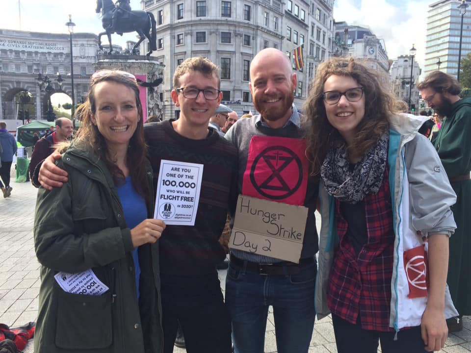 Picture shows Anna Hughes, Neil, Eddie and Helen at a Christian Climate Action event. They are smiling with their arms around each other and Anna is holding a Flight Free Pledge page in her hand. Eddie has a sign round his neck which reads 'hunger strike day 2'. There are people chatting behind them and they are outside. 