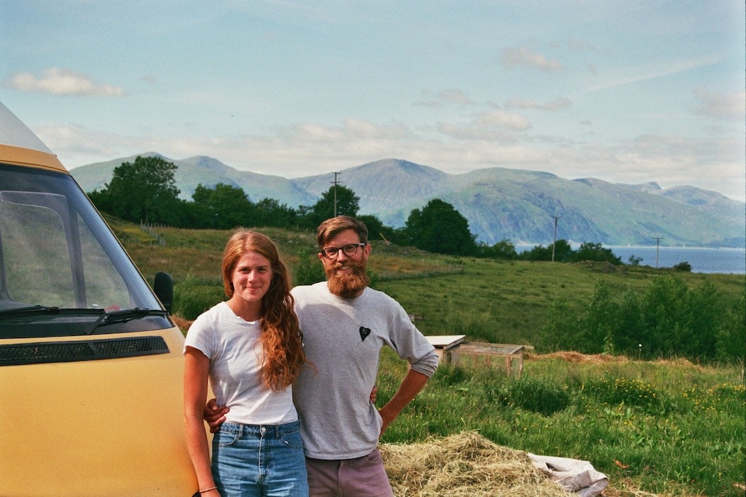 Picture shows Athlyn and Tim with their arms around each other. They are leaning on the hood of a yellow van. With grassy fields and far away mountains in the background. The picture was taken on a film camera. 
