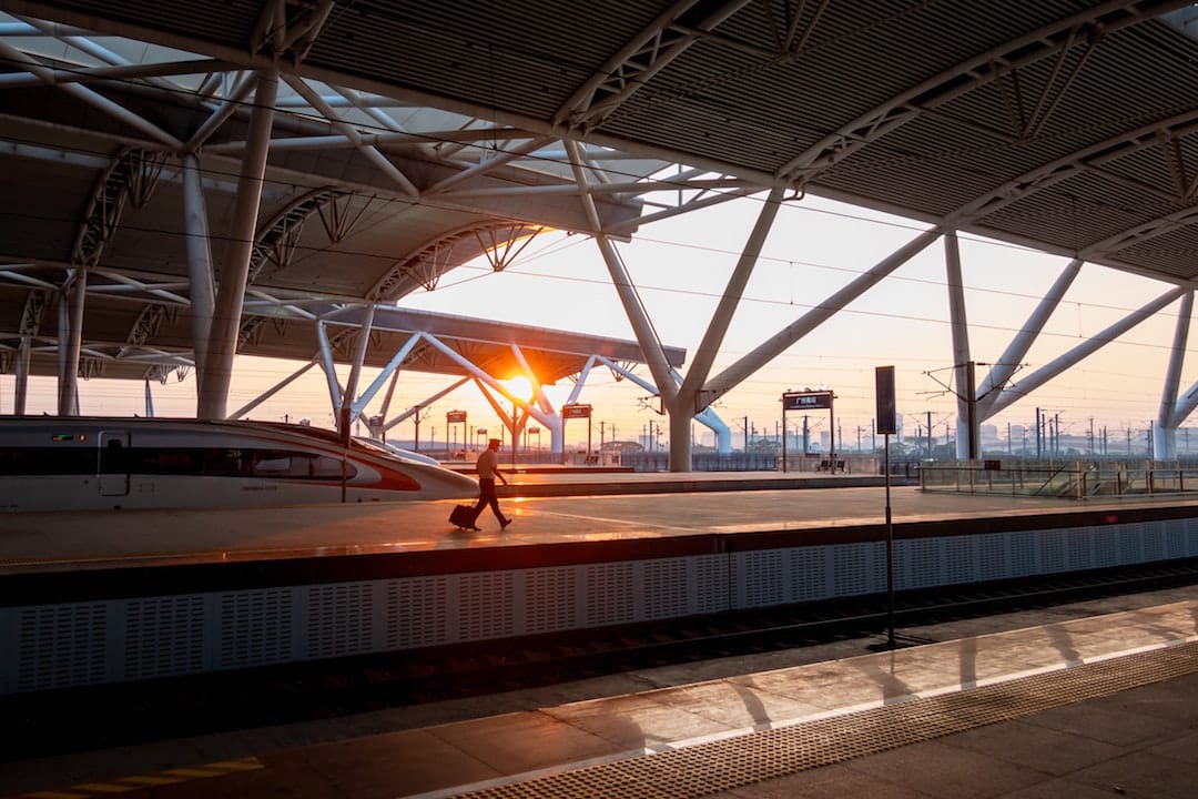 A passenger is walking across a rail platform in a Chinese train station. It is dusk and the sun is shining through the structure of the platform roof. 