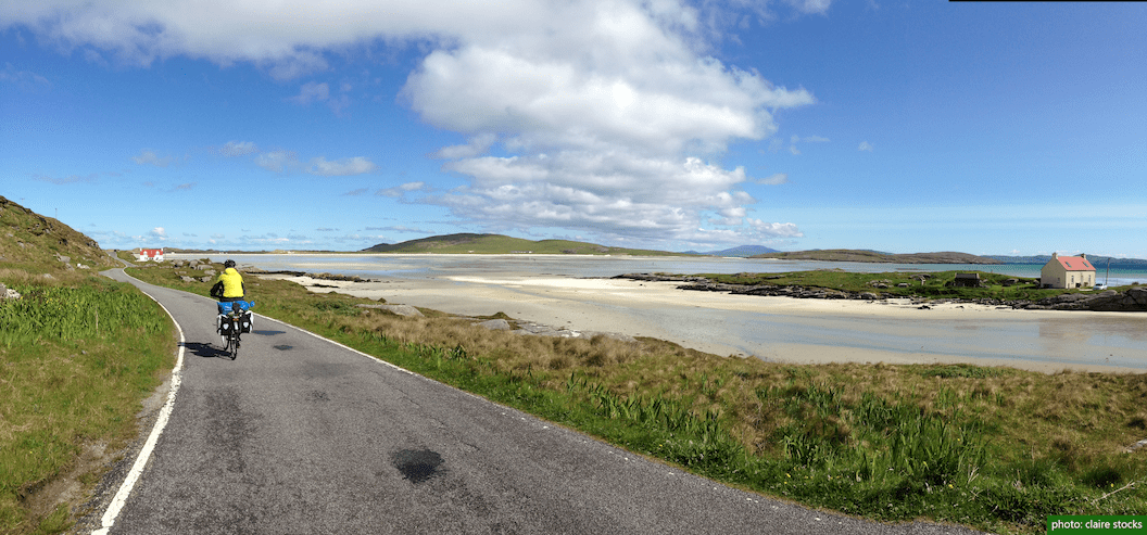 Picture shows a person cycling along a narrow road next to a white sandy estuary. In the distance is a small white house with a red roof and low rolling green hills. 