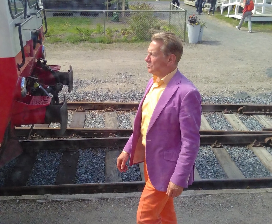 Picture shows Michael Portillo walking along next to train tracks. In the background are people walking in and out of a cafe. 