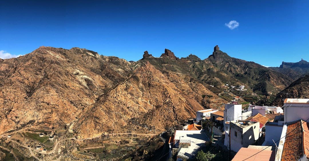 Picture shows a rocky mountain top in Gran Canaria. White and terracotta buildings line the right side of the frame. 