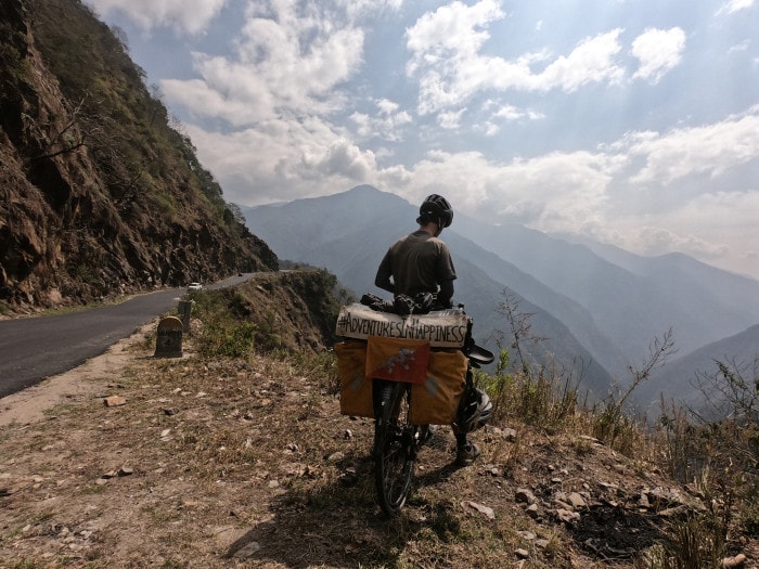 Picture shows man on bicycle parked at the side of a mountain road. He has his back to the camera and is looking over the edge. In the distance are mountain tops and rolling clouds. 
