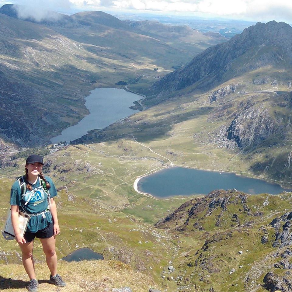 Picture shows Helena at the top of a peak in Snowdonia. Behind her is a steep decline into a grassy valley with small lakes. Part of the valley is in bright sunshine and part of it is in shade. 