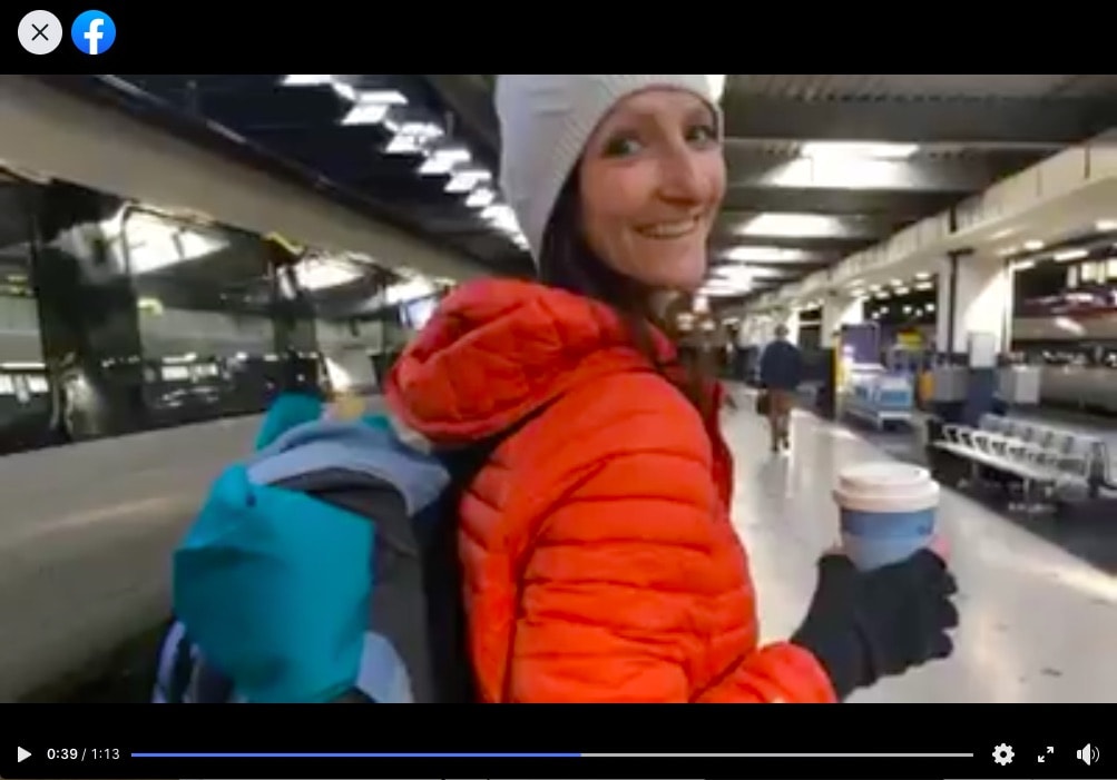 The image is a screenshot from the video on facebook mentioned below. Anna Hughes is turning around to the camera and smiling, wearing an orange puffer coat and carrying a cup of coffee. She is in a station. 