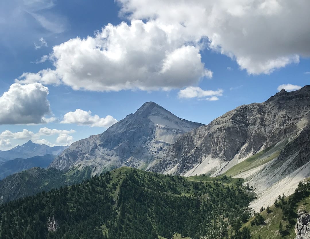 Image shows Oulx in the Alps. It is taken in the summer so there is no snow on the peak, and there is lots of green vegetation in the foreground. The sky is very blue with white fluffy clouds.