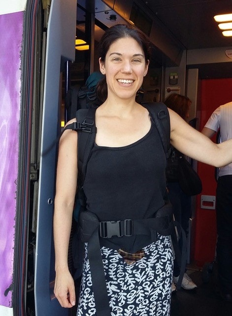 Milena is standing in the doorway of a train smiling and wearing a large rucksack. 