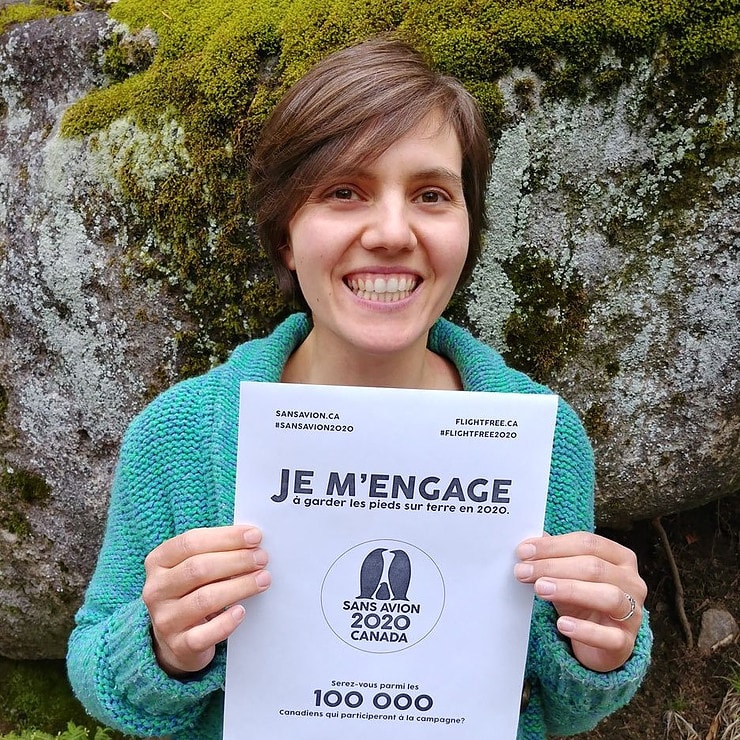 Nathalie holds a piece of paper with the Flight Free Canada 2020 pledge on it written in French. She is wearing a turquoise jumper and is smiling widely. 