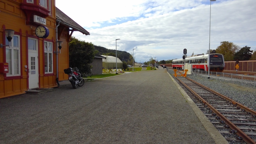The picture shows a train travelling along tracks next to a small station. There are some trees in the background. The station is red brick and it is an overcast day. 