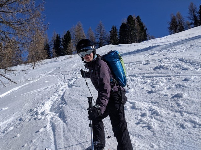 Picture shows skier facing the camera. She is wearing full ski gear including mirrored sunglasses and helmet. Behind them is and expanse of white snow with pine trees in the background. The sky is deep blue. 