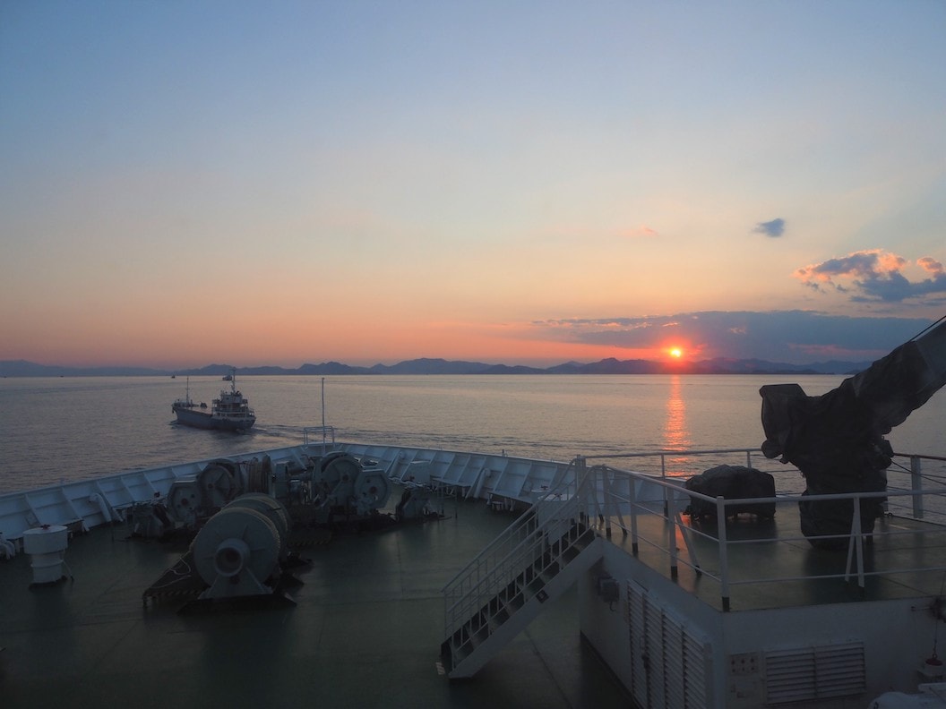 Picture shows the end of the ferry. Beyond the railings you can see the sea with low craggy hills in the distance. The sun is setting and the sky is orange yellow and blue. 