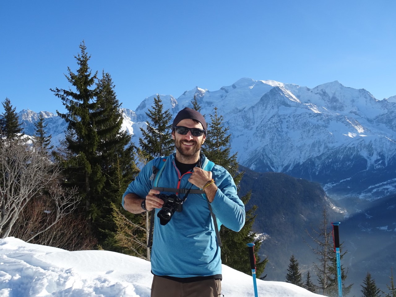 Josh stands on the side of a snowy mountain, with pine trees behind him and a clear view of a neighbouring peak. The sky is bright blue and it matches the colour of Josh's blue fleece. 