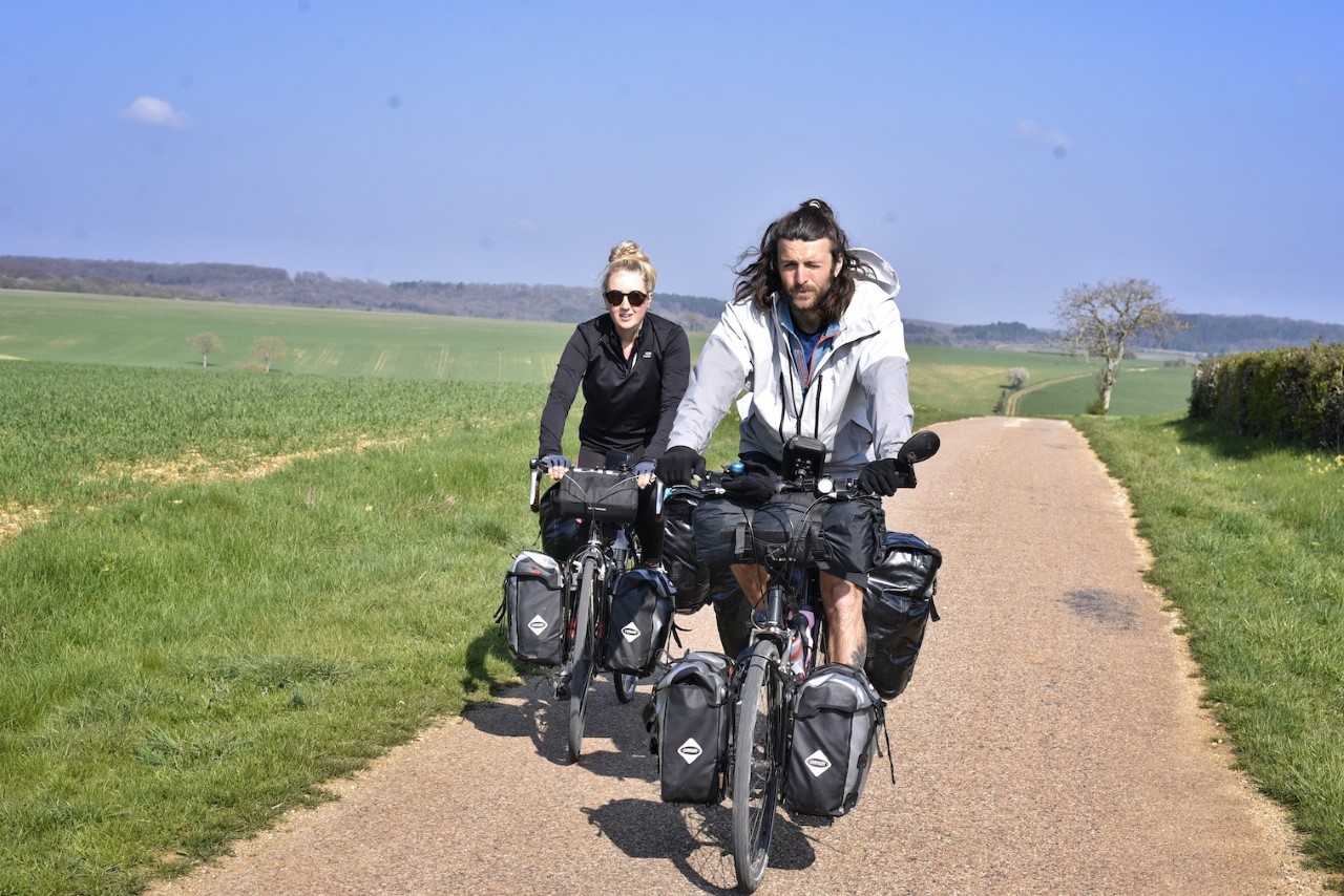 Josh and Sarah are on bikes with many panniers. It is a sunny day and they are on a pale cycle track in the middle of large rolling green fields