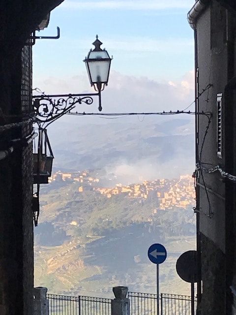 Picture shows the view down a street between two buildings. Railings at the end of the street protect from the sheer drop. In the distance are foggy green hills with houses clustered on them and rolling clouds further back. The town is in the sun and the close up buildings are silhouetted in contrast to the bright view. 